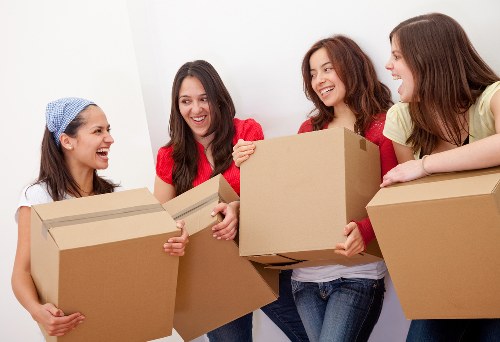 Four women with light to medium skin tones in casual clothing, including a woman wearing a blue headscarf, are standing indoors against a plain white wall. They are holding medium to large cardboard boxes, which are sealed and appear to contain household items. The woman on the far left is smiling and holding a box in both hands, while the second woman from the left, dressed in a red top, is smiling broadly and gripping a box with her right hand. The third woman, also smiling, is wearing a red jacket over a white shirt and is holding a box in her right hand, with her left hand resting against her side. The woman on the far right is wearing a light yellow top and has her left arm resting on a box placed on a surface. The scene suggests a friendly and organized home move or packing activity, likely part of a house removals service in or near Docklands, London, as indicated by the context of Docklands Man with Van services, with all four women engaged in handling packing boxes in a well-lit, tidy interior space.