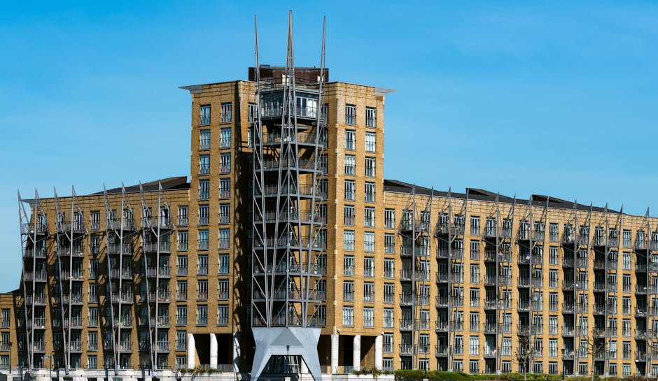 Photograph of a large modern residential building with multiple floors and numerous windows, situated in an urban area against a clear blue sky. The building features a central tower section with meta