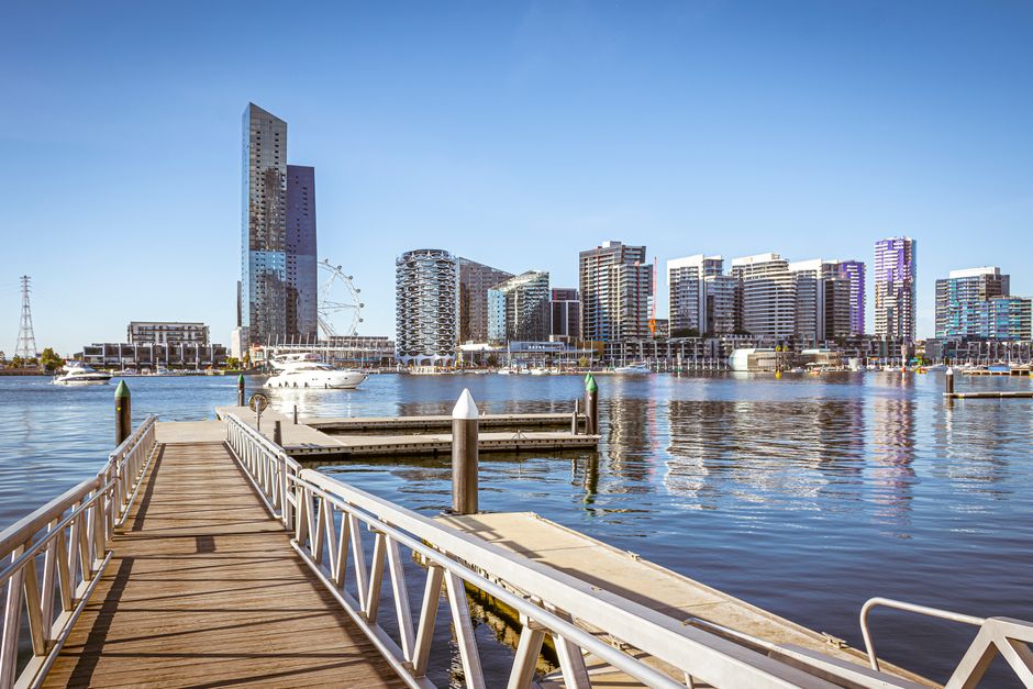 A view of a narrow waterway lined by modern multi-storey residential buildings made of yellowish brick and concrete, with black-framed windows and balconies, situated in Docklands. On the left side, c
